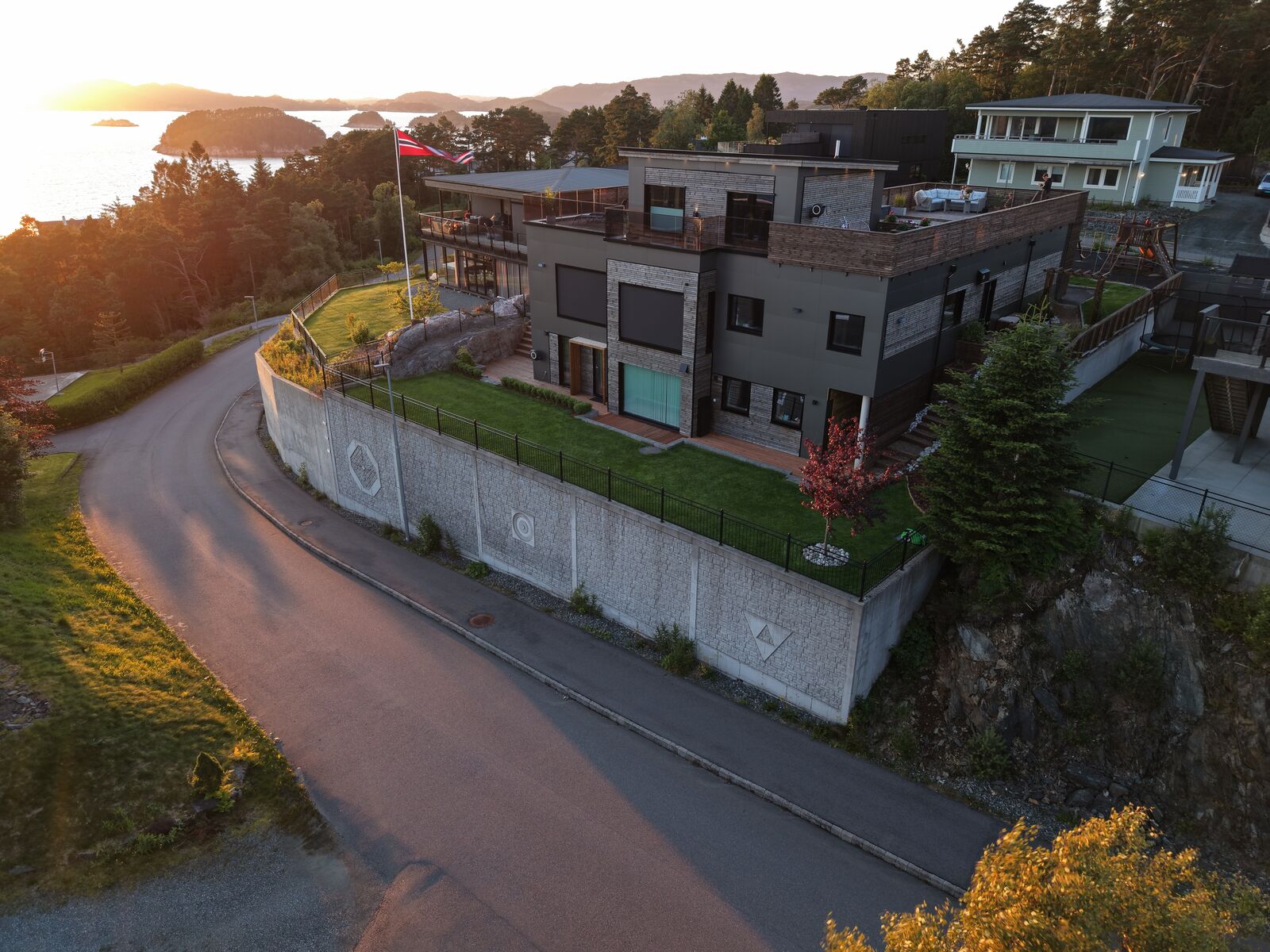 Dronefoto av eiendommen sett fra veien, med støttemuren og fjorden i bakgrunnen